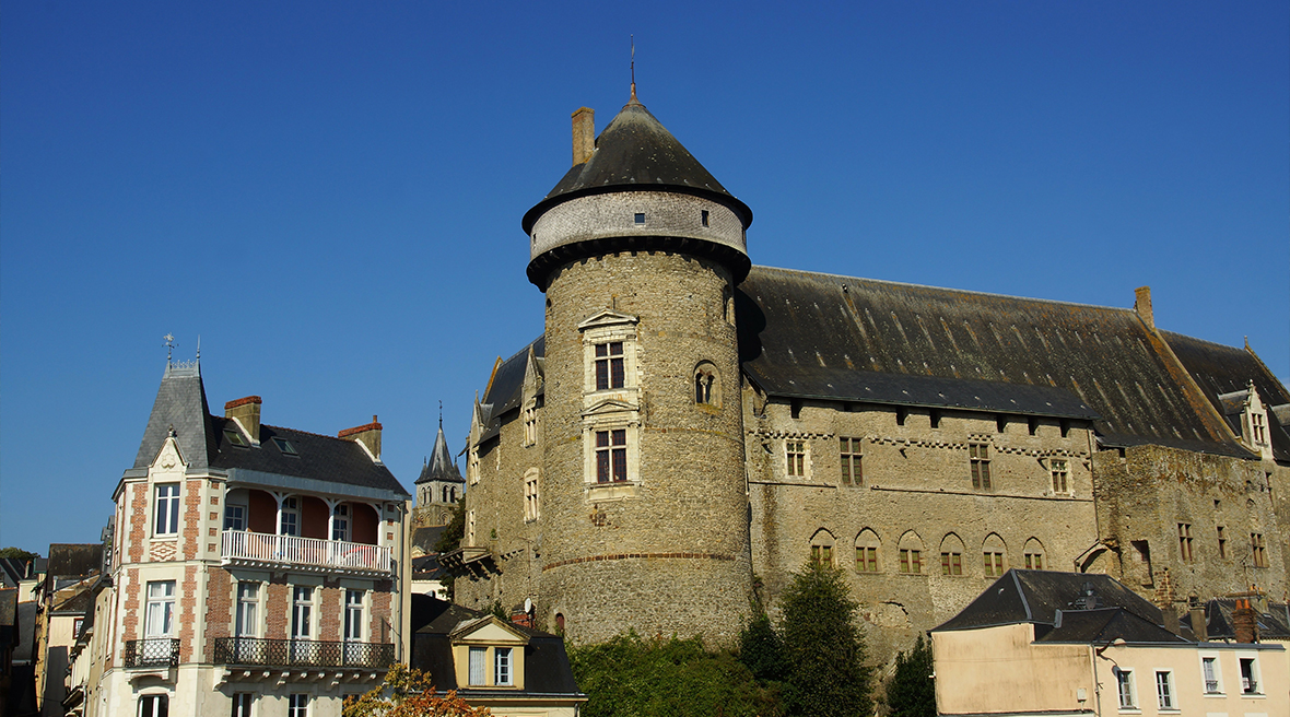 Large stone castle with a large round tower surrounded by ornate buildings under blue skies